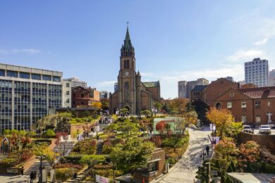 Myeongdong Cathedral in Seoul, South Korea, its Gothic-revival brick spire rising above surrounding city buildings, with autumn trees and a stone stairway leading up to the entrance.