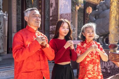 An Asian family father mother and young daughter dressed in bright cheongsams stands together, hands clasped in prayer, in a vibrant Chinese temple during Lunar New Year celebrations