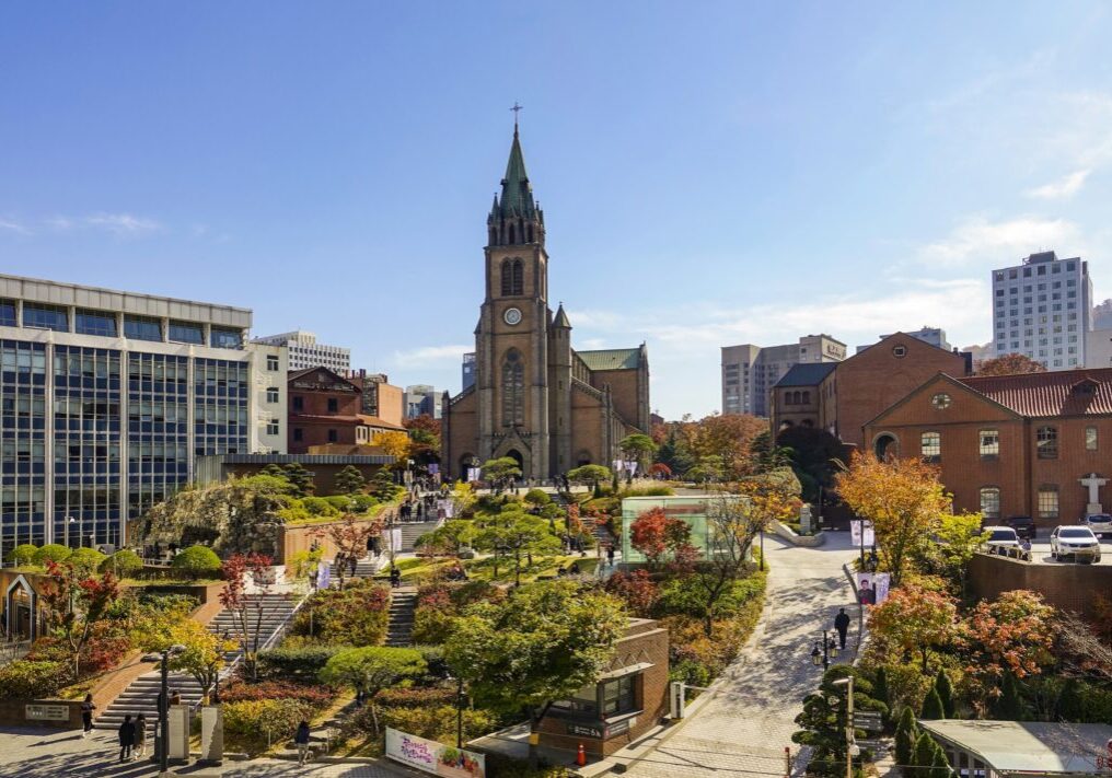 Myeongdong Cathedral in Seoul, South Korea, its Gothic-revival brick spire rising above surrounding city buildings, with autumn trees and a stone stairway leading up to the entrance.