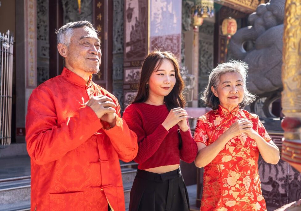 An Asian family father mother and young daughter dressed in bright cheongsams stands together, hands clasped in prayer, in a vibrant Chinese temple during Lunar New Year celebrations
